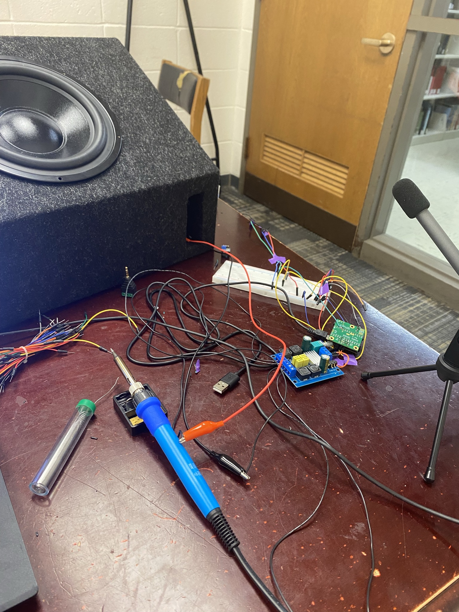 Electronics workbench with a subwoofer, breadboard circuits, amplifier boards, soldering iron, and microphone